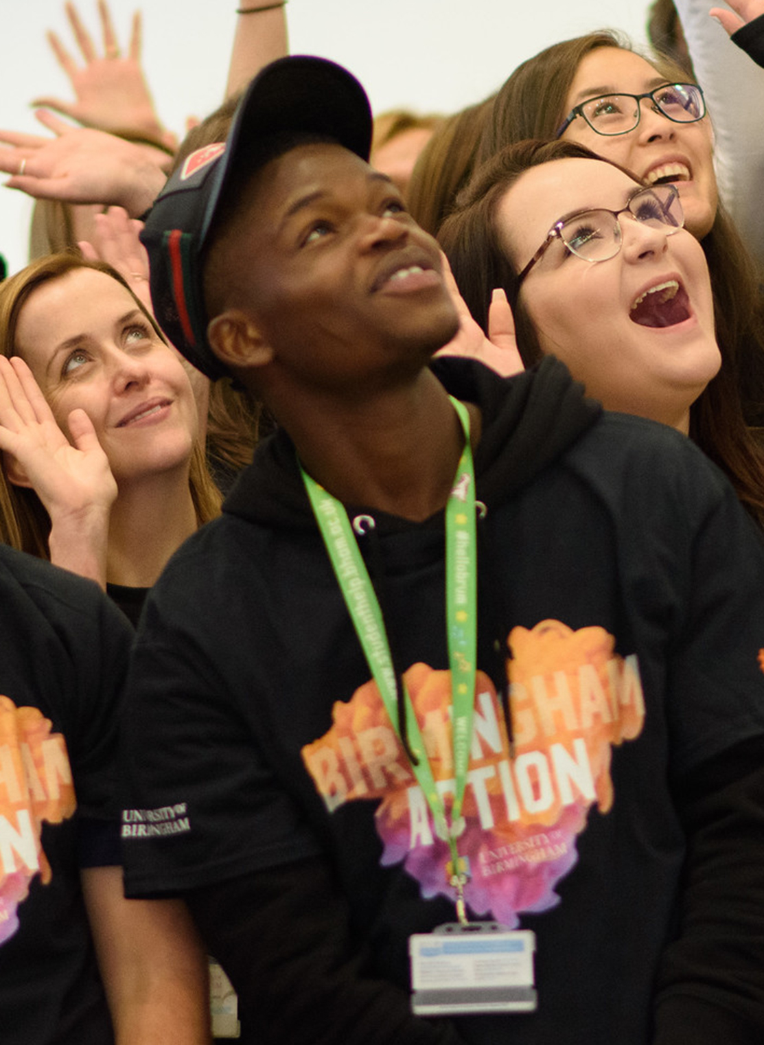 Male student looking up, wearing black cap, green lanyard and black, orange and purple t-shirt that says 'Birmingham in Action.'