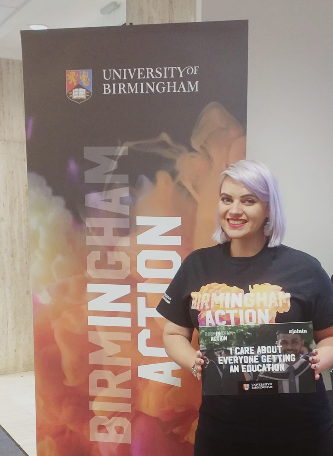 Female wearing black, orange and purple t-shirt that says 'Birmingham In Action' holding a sign that says 'I care about everyone getting an education' in front of a banner that says 'Birmingham In Action.'