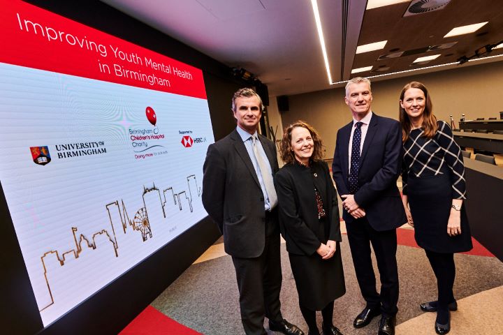 Two men and two women stand in front of a sign with University of Birmingham, HSBC and BCH logos on it