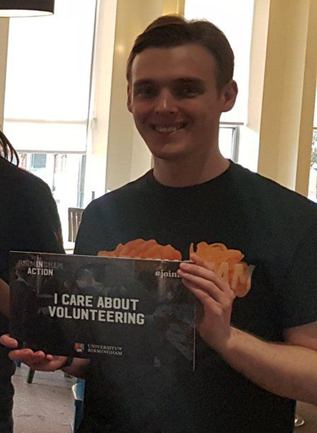 Male wearing black and orange t-shirt and holding a board that says ' I care about volunteering'