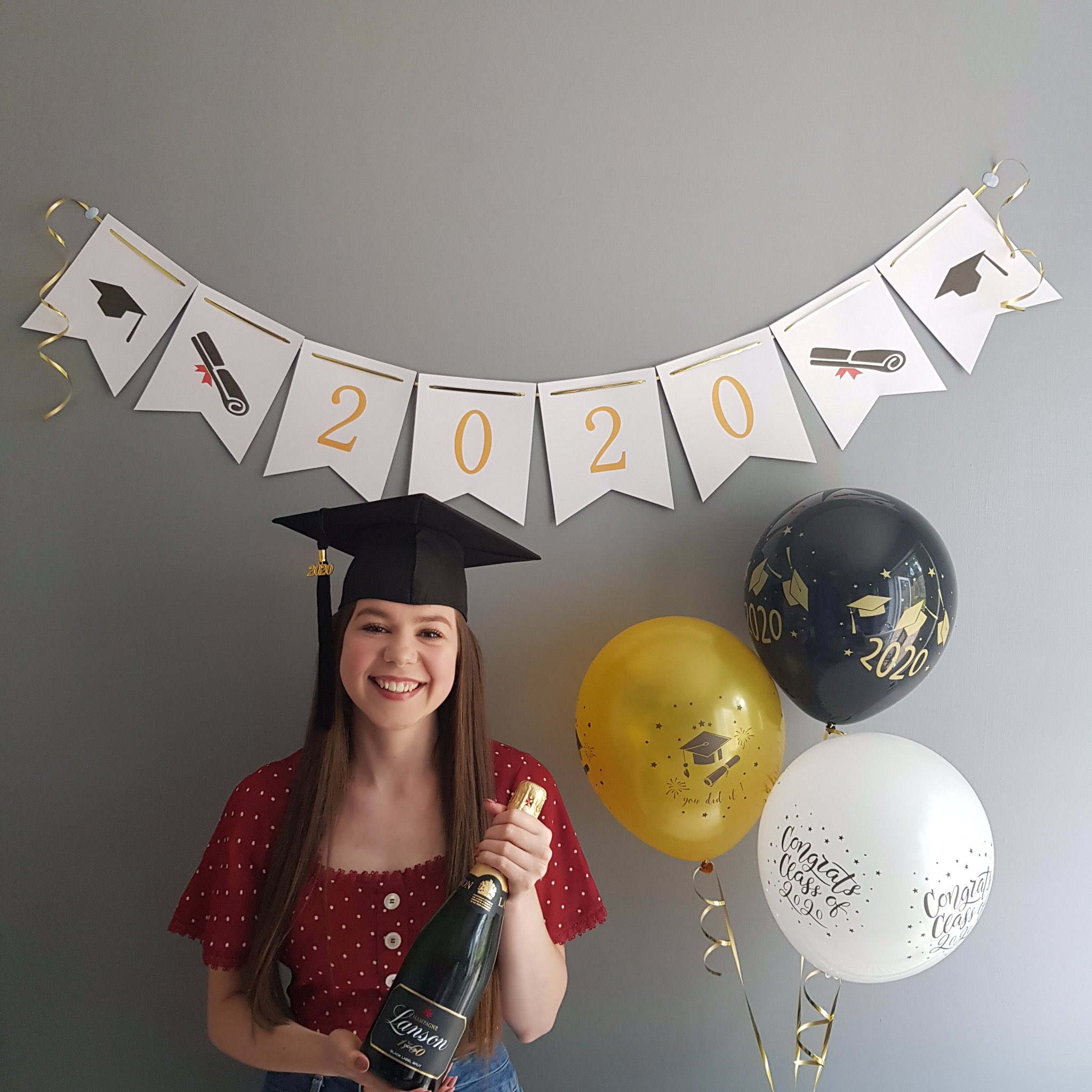 Girl smiling wearing graduation cap with champagne in hand, banner behind her that says 2020 and three balloons next to her