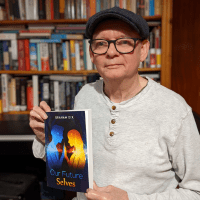 Graham wears a flat cap and glasses, and holds his book 'Our Future Selves' to show the camera. 