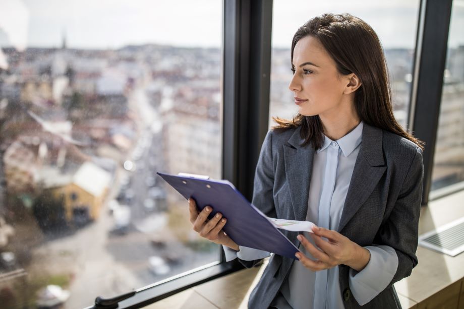 Young female lawyer looks out a window over buildings, while holding a clipboard