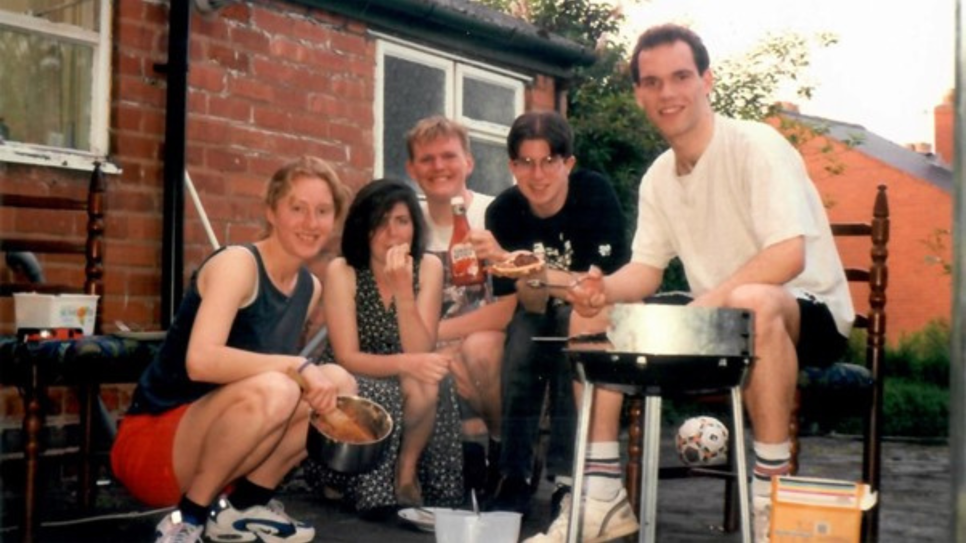 Group of five friends gathered outdoors near a brick house, enjoying a casual barbecue with sauce and food in hand.