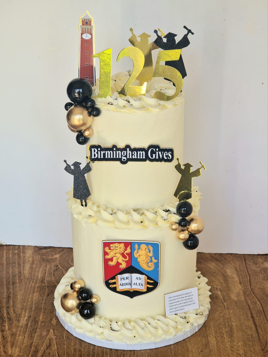 Two-tiered cream cake decorated with gold and black balloons, graduation cap silhouettes, and a red clock tower topper celebrating 125 years of Birmingham Gives.