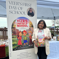 Gusharan smiles at the cameraholding a copy of her children's book. The book has a brightly coloured illustration of three children walking to school. 