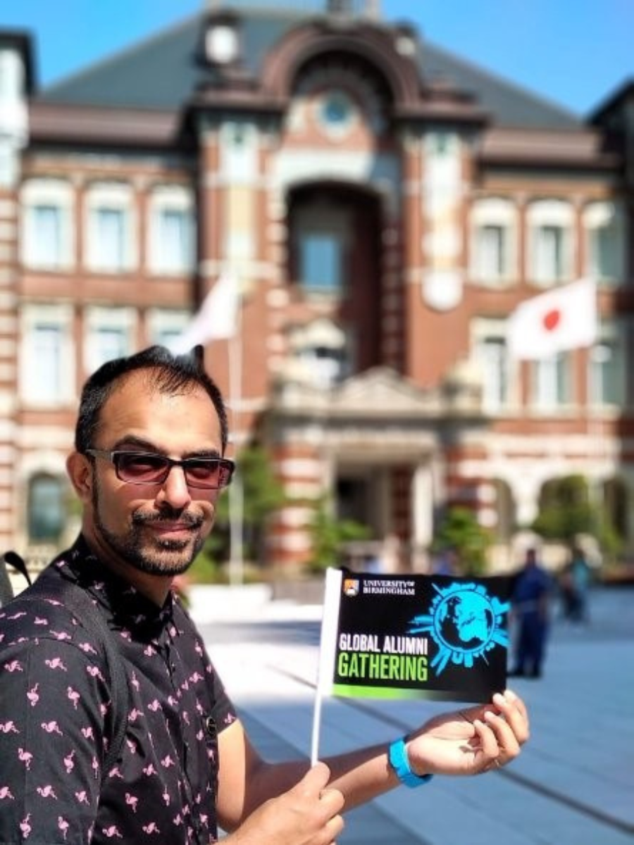Person holding a University of Birmingham Global Alumni Gathering flag in front of a historic red brick building with Japanese flags in the background.