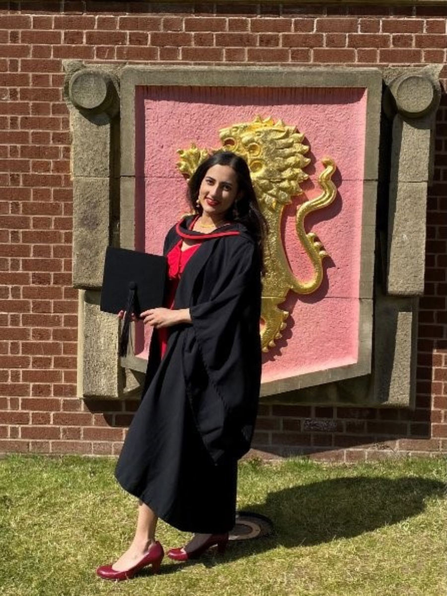 Graduate in black gown and red dress holding a diploma, standing in front of a brick wall with a golden lion emblem on a pink background.